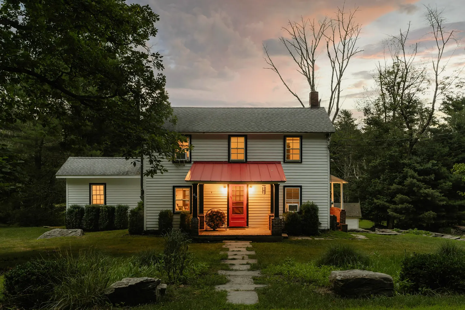 A small white house at dusk with its windows lit and a red awning over the front door.