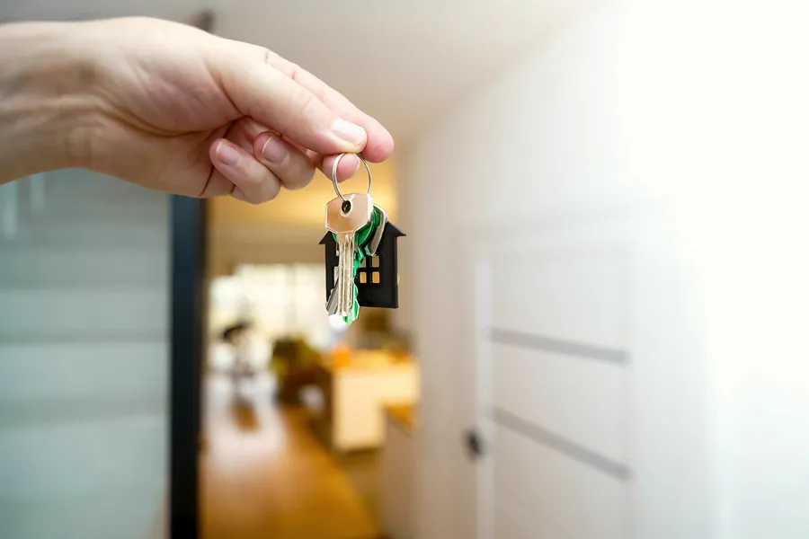 A hand holding a set of house keys on a little house-shaped keyring in a bright hallway.